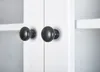 A close-up view of two black knobs on a white cabinet door. Produktbild-Vorschau
