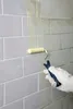 A hand wearing a white glove is using a paint roller to apply a yellow primer to a tiled wall. Produktbild-Vorschau