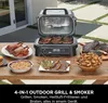 An outdoor grill and smoker is set up on a wooden table, surrounded by various cooked foods like steaks, chicken, and vegetables. Voir les images du produit