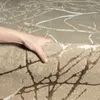 A hand is shown lifting a corner of a beige and gray rug with a branch-like pattern. Produktbild-Vorschau