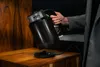 A hand holding a black electric kettle over its charging base on a wooden surface. Podgląd zdjęcia produktu