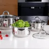 A set of stainless steel pots with glass lids on a white countertop, with a kitchen appliance in the background. Some cherry tomatoes and lettuce are also visible. Náhled obrázku produktu