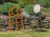A rusty iron window frame and a black metal chair are placed on a grassy area in front of a garden. Produktbild-Vorschau
