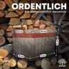 A wicker basket filled with firewood, placed in front of a stack of logs and a wooden cart wheel. Produktbild-Vorschau