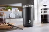 A black electric coffee grinder is shown on a kitchen counter next to a French press and a jar of coffee beans. Produktbild-Vorschau
