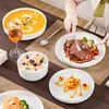 A table setting with various dishes, including soup, steak, fruit salad, and pasta, with a person's hands visible preparing to eat. Produktbild-Vorschau