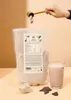 A hand is adding protein powder from a scoop into a large bag of protein powder. A glass of chocolate protein shake and some chocolate pieces are on the table. Produktbild-Vorschau