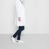 A person wearing a white lab coat and dark jeans walks to the right, showing off white shoes. Produktbild-Vorschau