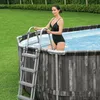 A woman in a black swimsuit is standing on the ladder of an oval above-ground swimming pool in a backyard. Náhled obrázku produktu