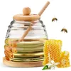A glass honey jar with a wooden lid and dipper, accompanied by honeycomb and bees. Produktbild-Vorschau