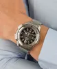A person wearing a light blue collared shirt is showing off a silver and black wristwatch on their left wrist. Produktbild-Vorschau