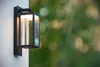 A modern outdoor wall light fixture mounted on a textured white wall, with a blurred green background of foliage. Podgląd zdjęcia produktu
