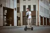 A man rides an electric scooter down a city street, with modern buildings in the background. Produktbild-Vorschau