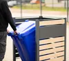 A person is placing a blue trash bin into a gray and wood-paneled trash bin enclosure. Náhled obrázku produktu