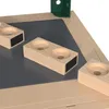 A close-up view of a toy workbench with several beige blocks with circular indentations and black rectangular inserts, placed on a gray surface. A green corner piece is visible in the upper right. Produktbild-Vorschau