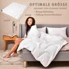 A woman is sitting on a bed wrapped in a white duvet, with a bedside table and lamp visible. Produktbild-Vorschau
