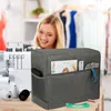 A gray sewing machine cover with storage pockets is shown in front of a sewing machine and a blurred woman. Produktbild-Vorschau