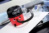 A small, red and black Henry vacuum cleaner is placed on a white surface, with a clear plastic container and other accessories in the background. Produktbild-Vorschau