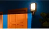 An outdoor wall light with a camera is mounted on a blue wall next to an orange door. A blue light beam emanates from the camera, indicating its field of view. Produktbild-Vorschau