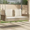 A beige rattan swing bench with cream cushions is shown outdoors on a wooden deck, with a wooden fence and lattice in the background. Produktbild-Vorschau