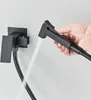 A hand is holding a black bidet sprayer, demonstrating its use with water spraying from the nozzle. A black bidet faucet and holder are mounted on a white wall in the background. Náhľad obrázku produktu