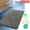 A dark gray bath mat with a leaf pattern is shown on a tiled floor next to a cabinet. Several marketing logos and text are overlaid on the right side of the image. Produktbild-Vorschau
