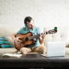 A man is sitting on a couch and playing a guitar while wearing headphones. A laptop and a notebook are on the coffee table in front of him. Náhled obrázku produktu