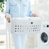 A person is holding a laundry basket filled with white clothes in a laundry room. Produktbild-Vorschau