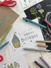 A collection of art supplies and birthday cards arranged on a wooden surface. Voir les images du produit