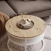 A round coffee table with a wood-look top and a white base, placed on a rug next to a sofa. Náhľad obrázku produktu