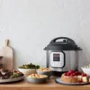 An Instant Pot Duo 7-in-1 Smart Cooker is displayed on a kitchen counter with various dishes, including mac and cheese, roasted vegetables, and a cheesecake, suggesting a meal preparation scenario. Podgląd zdjęcia produktu