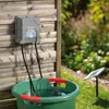 A solar-powered garden watering system is shown mounted on a wooden fence, with hoses leading to a green basin filled with water. A small solar panel is visible in the background. Produktbild-Vorschau