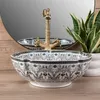 A close-up shot of a decorative basin sink with a gold faucet, set against a blurred background of a mirror and a plant. Anteprima dell’immagine prodotto