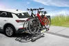 A white car with a bicycle rack attached to the rear, carrying two bicycles, parked on a paved surface near a grassy area and the ocean. Anteprima dell’immagine prodotto
