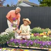 A couple is gardening in front of a dark gray fence, surrounded by colorful flowers. Produktbild-Vorschau