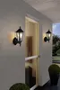 Two outdoor wall lamps are mounted on a textured wall next to a glass door. A potted plant is visible in the foreground and background. Anteprima dell’immagine prodotto