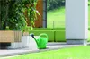 A green watering can sits on a concrete patio next to a planter with lush green foliage. In the background, there are large green cushions and a wooden structure. Náhľad obrázku produktu