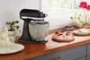 A black KitchenAid stand mixer with a decorative ceramic bowl sits on a wooden countertop next to a pitcher of lemonade and some desserts. Produktbild-Vorschau