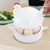 A white chicken-shaped egg cooker is placed on a wooden table, with eggs inside the base. In the background, a plate of green salad and some yellow and orange food items are visible. Produktbild-Vorschau