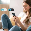 Zwei Webcam-Abdeckungen, eine weiße und eine schwarze, beide mit blauen Akzenten, überlagert auf einem Bild einer Frau, die ein Smartphone benutzt Produktbild-Vorschau