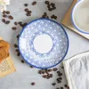 A close-up overhead view of a blue and white patterned saucer with coffee beans scattered around it. Produktbild-Vorschau