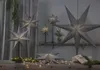 A collection of gray star-shaped lamps are displayed in a dimly lit room, some on stands and one hanging, with decorative lights and greenery. Podgląd zdjęcia produktu