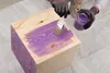 A person is painting a wooden cube with purple paint. Voir les images du produit