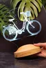 A 3D LED lamp shaped like a bicycle is displayed on a wooden base, with large green leaves in the background. Produktbild-Vorschau