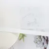 A close-up view of a white textured object, possibly a vase or a lampshade, with a sheer white curtain in the foreground. Behind the curtain, there are faint botanical illustrations and text. Produktbild-Vorschau