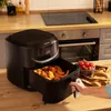 A black air fryer is on a wooden countertop in a kitchen, with its drawer pulled out to reveal fried food. A bowl of cherry tomatoes and a napkin are nearby. A person's hand is visible pulling the drawer. Produktbild-Vorschau