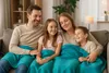 A family of four is sitting on a couch, covered by a teal blanket, and smiling. Produktbild-Vorschau