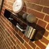 A black shelf mounted on a brick wall, holding CDs and decorative balls, with a clock above. Produktbild-Vorschau