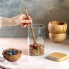 A hand is shown stirring granola in a glass jar with a wooden spoon. A bowl of granola with blueberries and yogurt is in the foreground, and two wooden bowls are stacked in the background. A bamboo lid is on the right. Produktbild-Vorschau