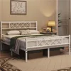 A bedroom scene featuring a white metal bed frame with a diamond pattern headboard and footboard, dressed with bedding and a green throw blanket. A bedside table with a lamp and decorative items is visible to the right. Podgląd zdjęcia produktu